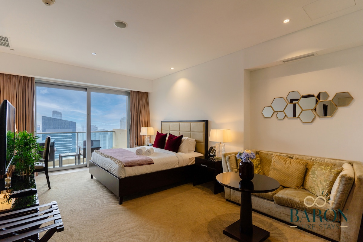 A welcoming studio apartment features a spacious bed with neatly arranged linens, paired with decorative pillows. A cozy seating area with a light-colored sofa is positioned next to a round table. Large windows provide ample natural light and showcase views of the vibrant city.