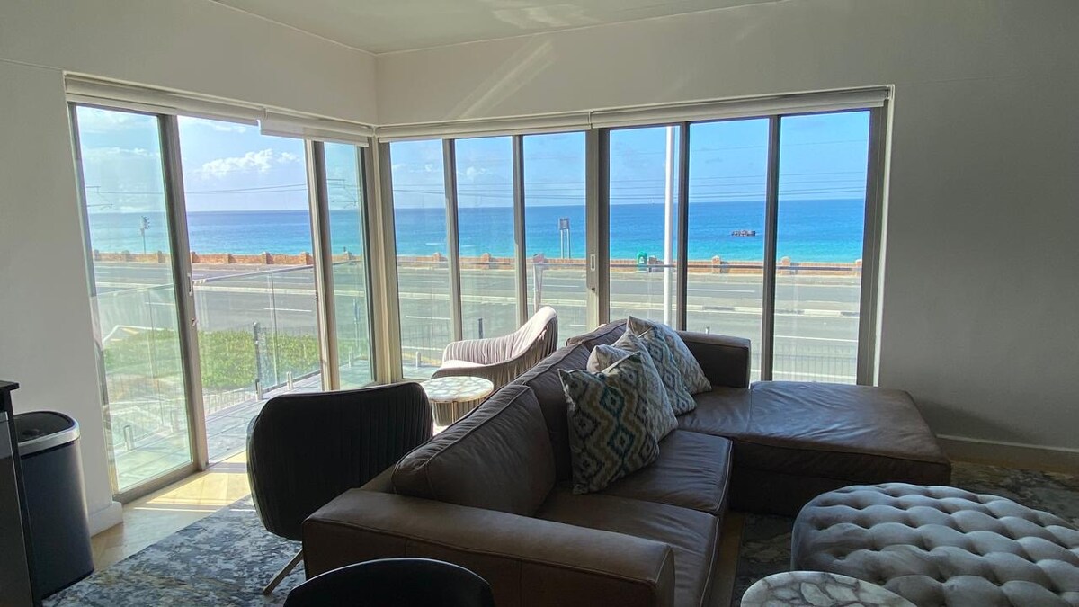 A spacious living area is highlighted by large glass windows offering unobstructed ocean views. Elegant furnishings include a modern sectional sofa, comfortable armchairs, and a textured coffee table. Natural light floods the space, enhancing the coastal ambiance.