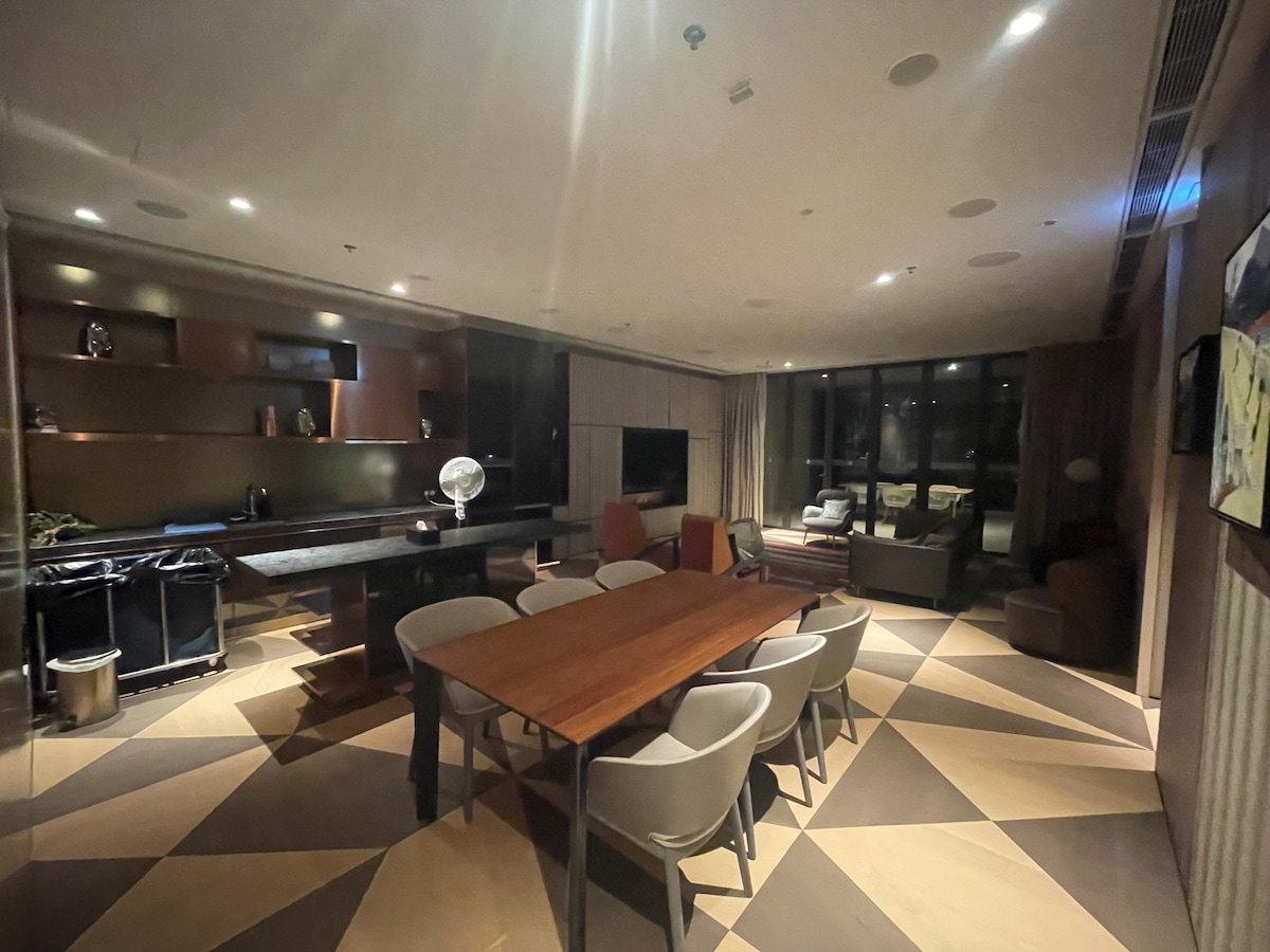 An open-plan living area features a long wooden dining table surrounded by several light-colored chairs. A sleek kitchen counter is visible in the background, with modern appliances and decorative elements displayed on the shelves. Floor-to-ceiling windows allow natural light to enter, enhancing the contemporary design.