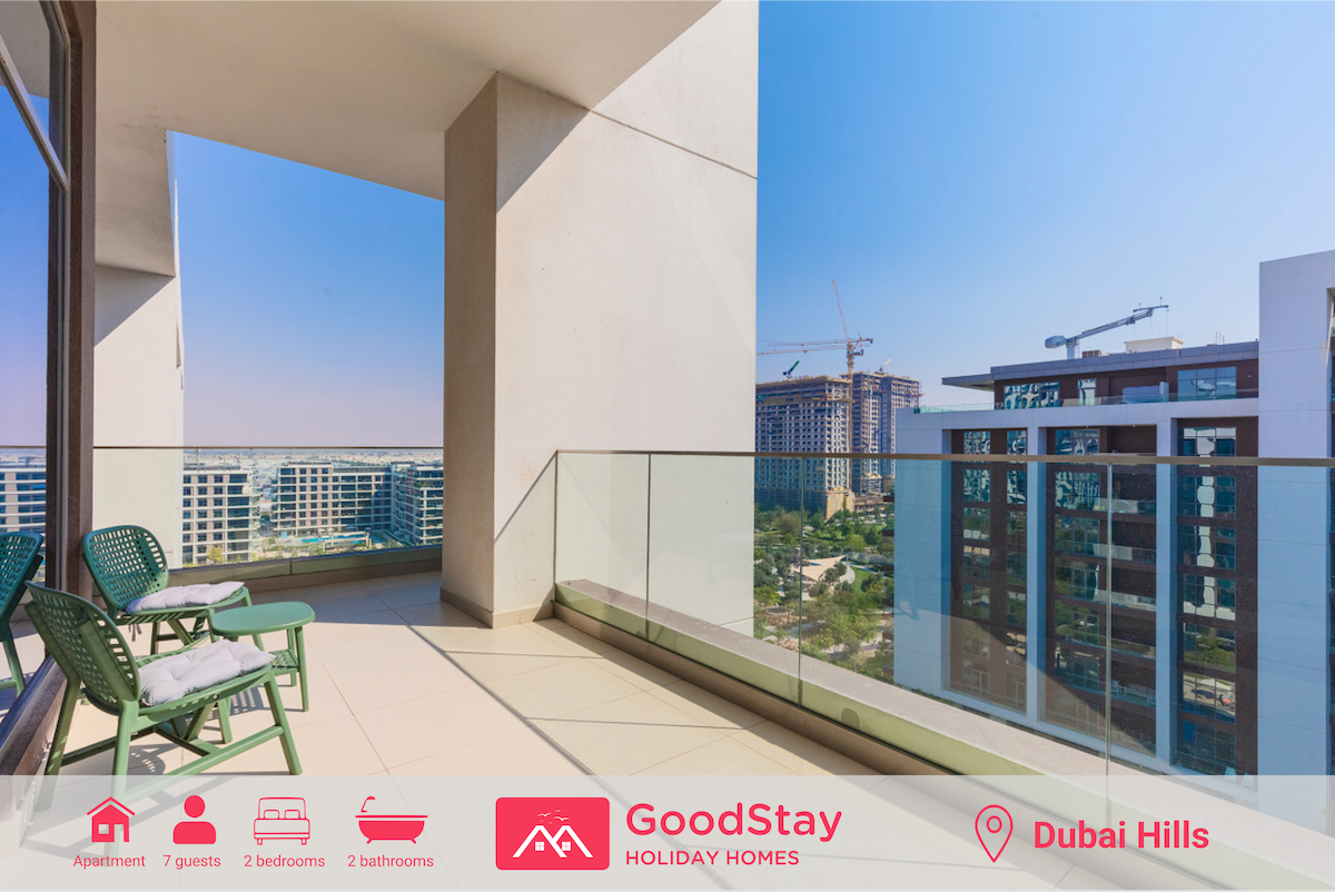A spacious balcony is featured, with views of the surrounding community and park. Two green chairs are positioned next to the glass railing, allowing for enjoyment of the scenery. The sleek, modern design is complemented by ample natural light flooding into the space.