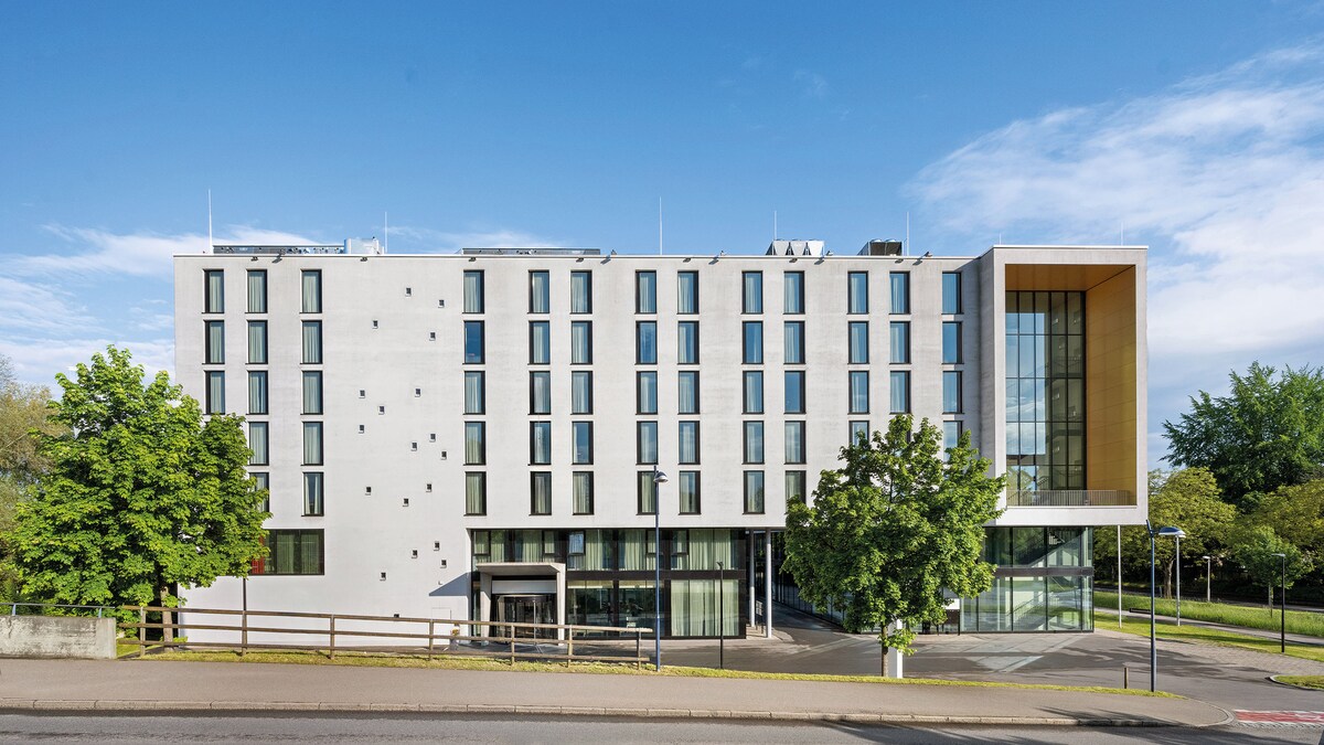 The hotel exterior features a modern architectural design with a facade comprised of light-colored panels and numerous windows. Large trees and landscaping are present at the front, contributing to a visually appealing entrance. A clear blue sky complements the contemporary structure.