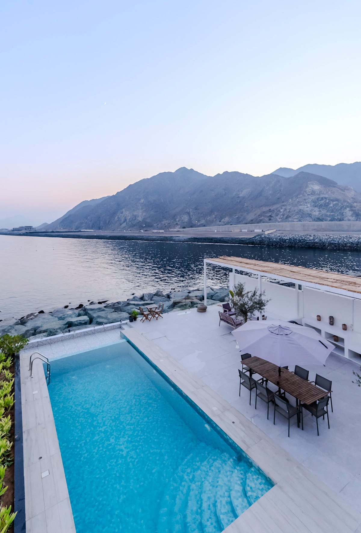 A serene outdoor area features a private swimming pool, bordered by a spacious terrace with a dining table and chairs, overlooking calm waters. Majestic mountains rise in the background, creating a picturesque setting for relaxation by the beach.
