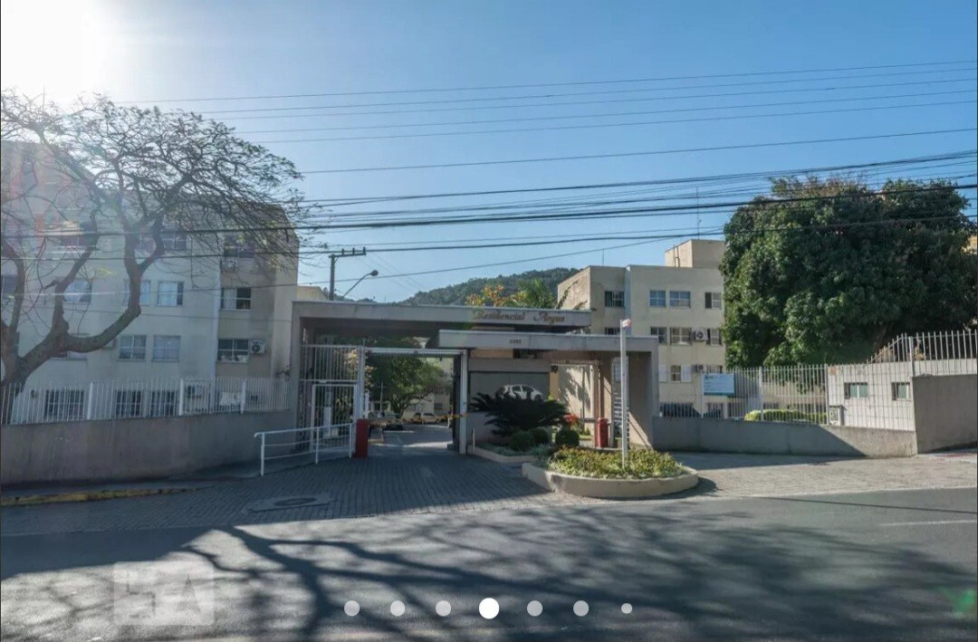 The entrance to the property is shown, featuring a gated access with a security checkpoint and landscaped greenery. Surrounding buildings are visible in the background, set against a clear blue sky, presenting a welcoming atmosphere.