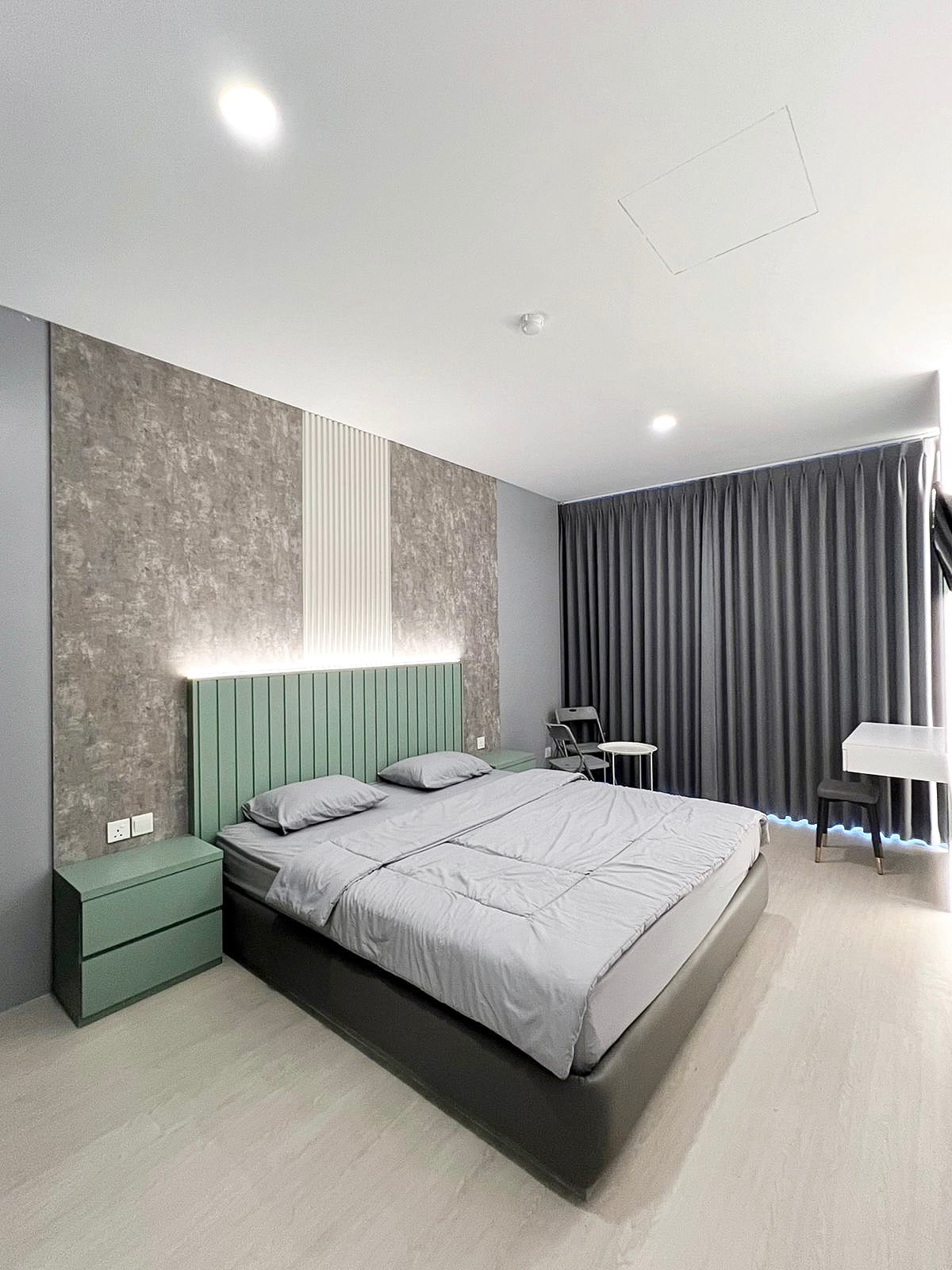 A spacious sleeping area is highlighted by a soft green headboard and neutral-toned bedding. Natural light filters through floor-to-ceiling curtains, creating a calming environment. A desk and chair are positioned nearby, providing a functional workspace.