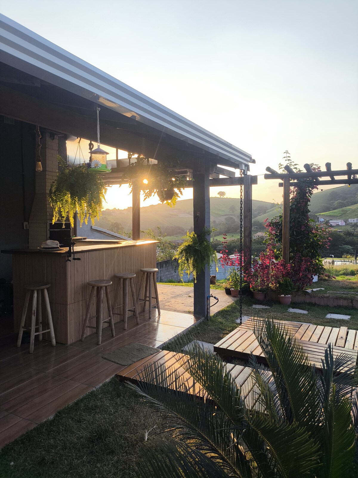 An open outdoor area features a wooden bar with stools, surrounded by lush greenery and hanging plants. Sunlight filters through the pergola, casting warm tones over the landscape beyond, which showcases rolling hills and vibrant flowers in the foreground.
