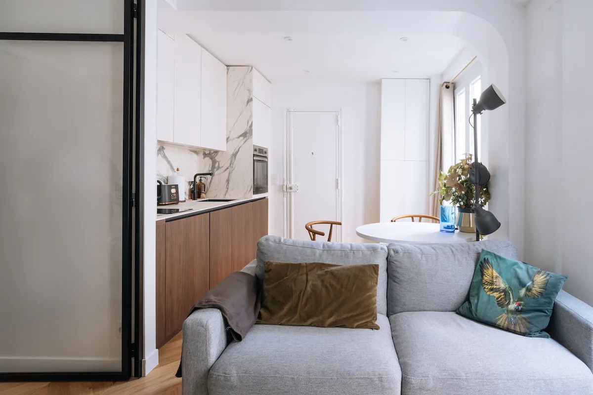A light-filled living area showcases a grey sofa adorned with decorative cushions. A round dining table is visible, accompanied by wooden chairs, while a modern kitchen with marble accents and wooden cabinetry is subtly integrated within the open layout.