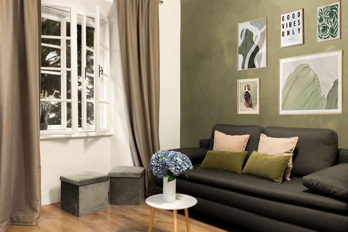 A modern living area features a dark sofa adorned with several cushions in neutral tones. A round coffee table with a vase of hydrangeas sits in front. Light filters through white window frames, which are paired with soft brown curtains, illuminating the textured green wall and art pieces.