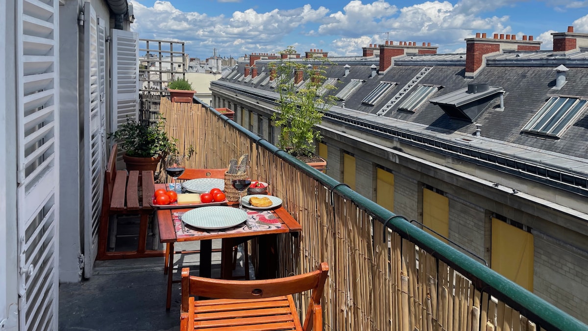 A balcony with wooden tables and chairs is displayed, set for a meal with dishes and fresh tomatoes. Potted plants add a touch of greenery. The view showcases rooftops under a partly cloudy sky, contributing to a serene outdoor dining ambiance.