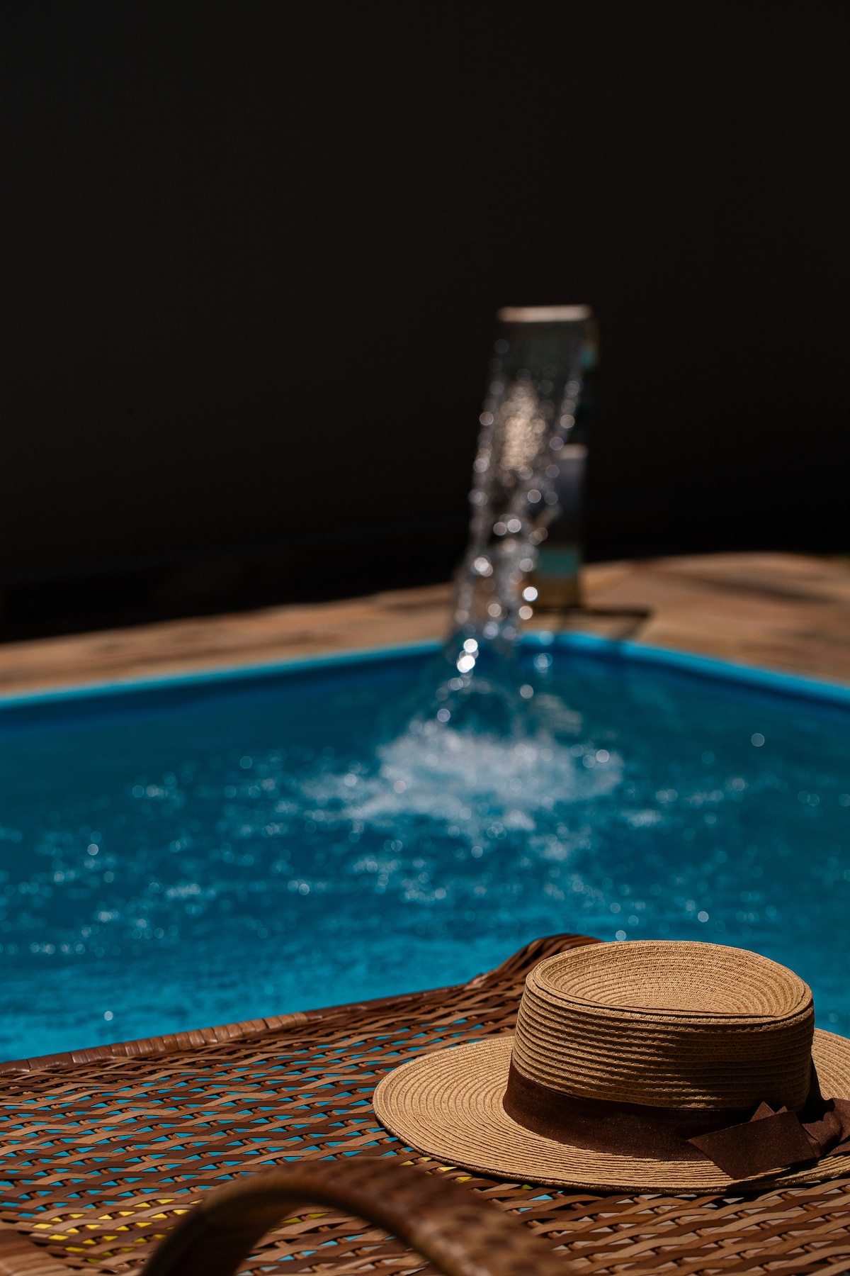 A refreshing swimming pool glistens under the light, featuring a water feature that creates gentle splashes. A straw hat is placed casually on the edge, complementing the woven texture of the surrounding lounge area.
