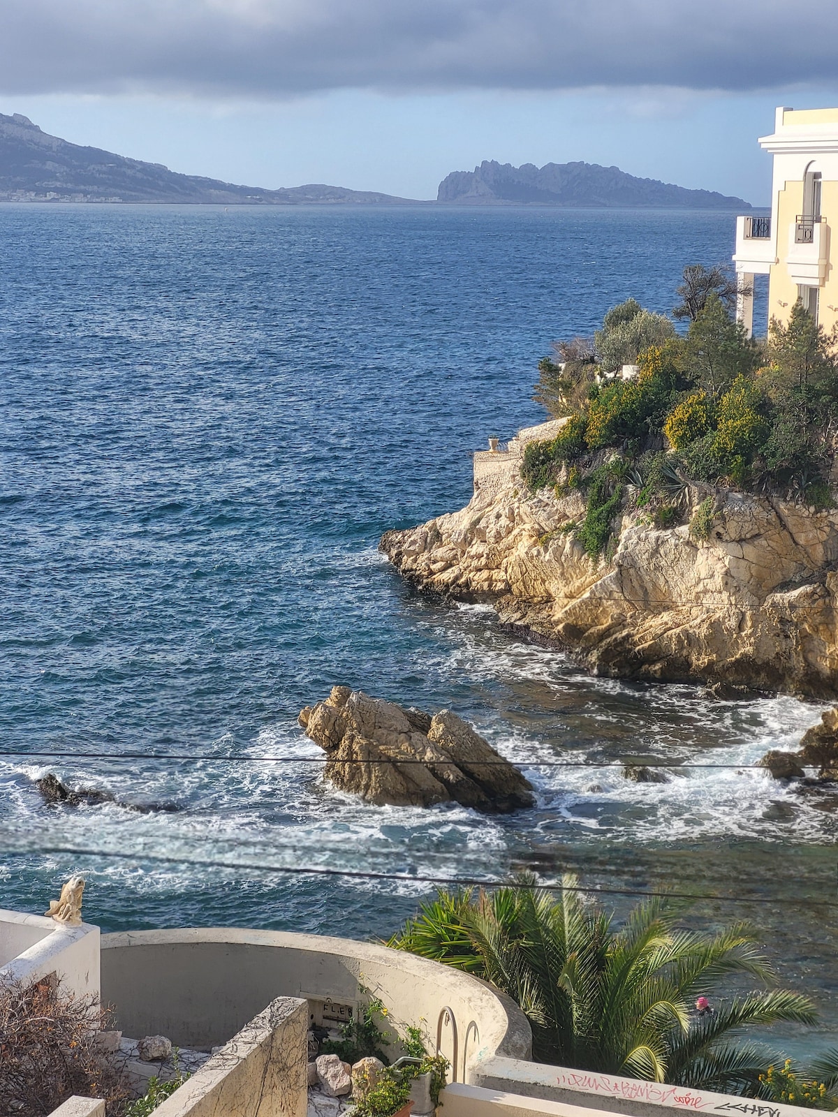 The image captures a rocky coastline where waves gently crash against the shore. Lush greenery clings to the cliffside, while distant hills rise above the shimmering blue waters. The sky is partly cloudy, creating a serene and inviting atmosphere by the coast.