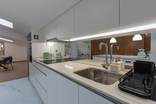 A modern kitchen area is presented, featuring sleek white cabinetry and a light countertop. Stainless steel sinks are positioned under a well-lit space, with minimalist kitchen appliances visible. The open layout allows for easy movement, complemented by natural light streaming through overhead skylights.