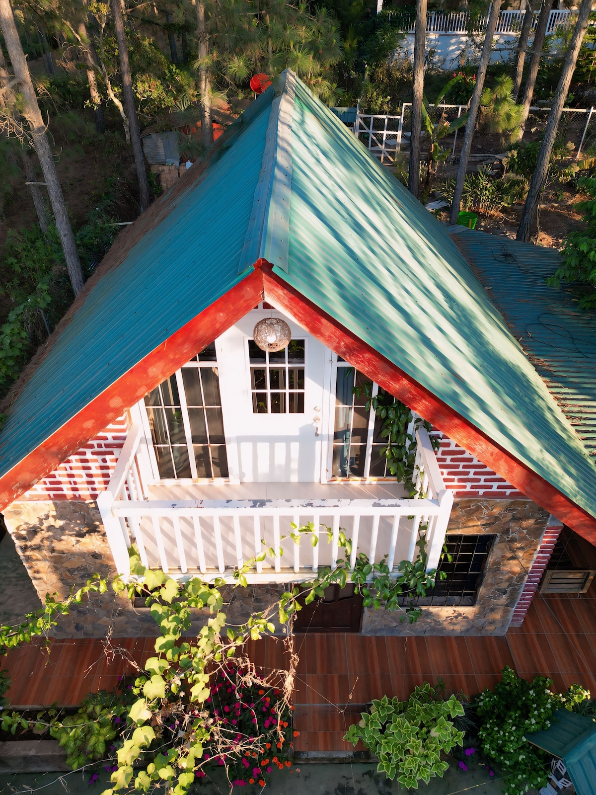 A bird's-eye view of a rustic cabin reveals a sloped metal roof with vibrant green hues. The structure features brick accents and a white balcony door, surrounded by lush trees and colorful flowering plants in the outdoor area.