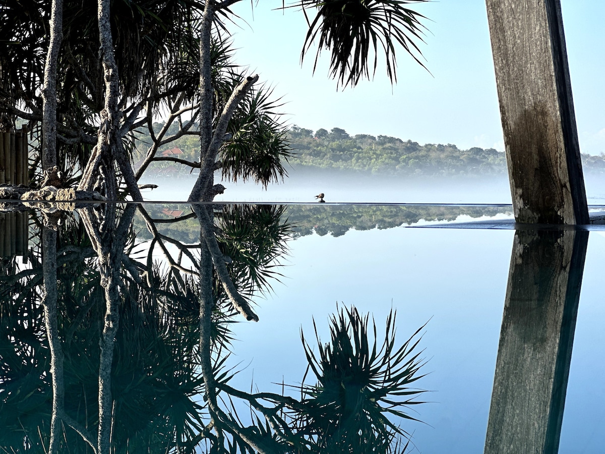 The image captures a tranquil pool reflecting the surrounding greenery and still water. Tropical plants and trees frame the view, adding to the serene atmosphere. A misty backdrop enhances the sense of calm, creating a connection with nature.
