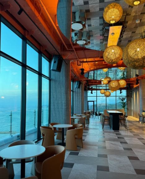 A spacious dining area features large windows offering views of the ocean. Pendant lights hang from a colorful ceiling, illuminating tables and chairs arranged for guests. A sleek bar area is visible in the background, complemented by a modern flooring design.