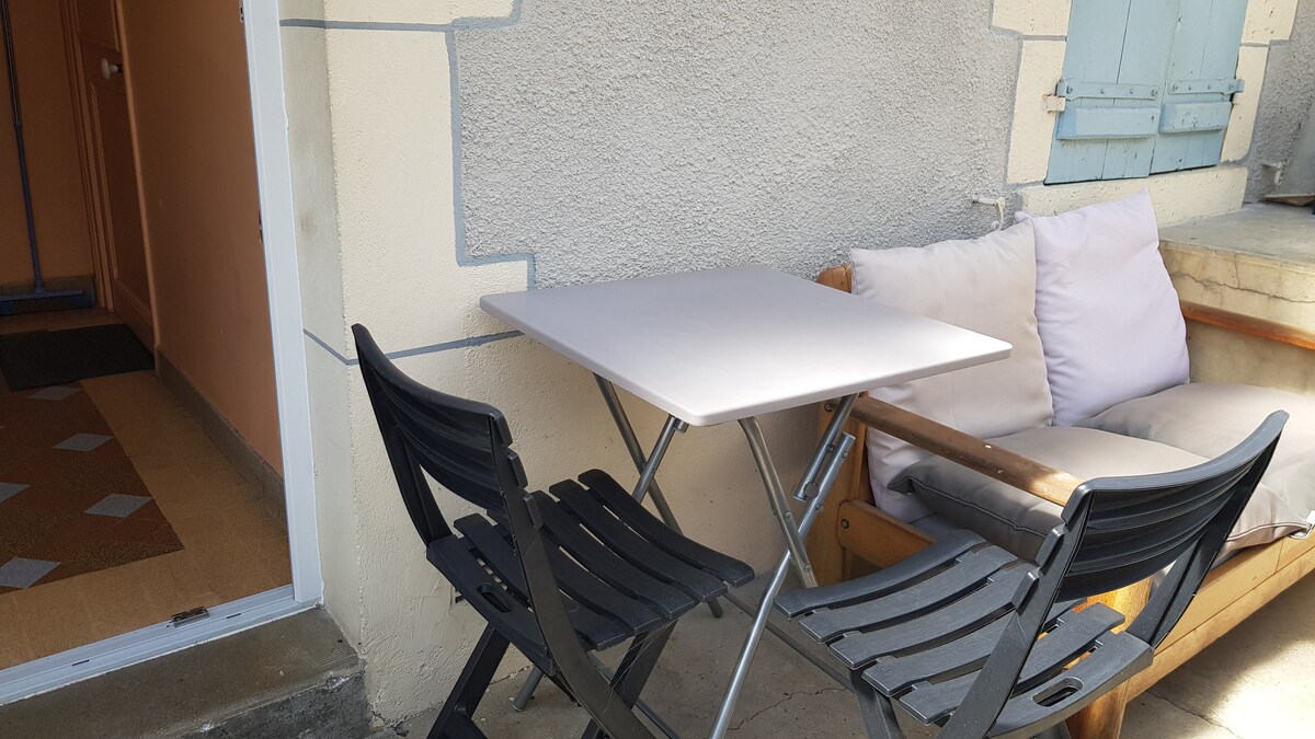 A small outdoor seating area features a foldable table paired with two black chairs. A comfortable wooden bench with soft cushions is visible nearby. The entrance to the studio is partially seen in the background, framed by light-colored walls and blue shutters.