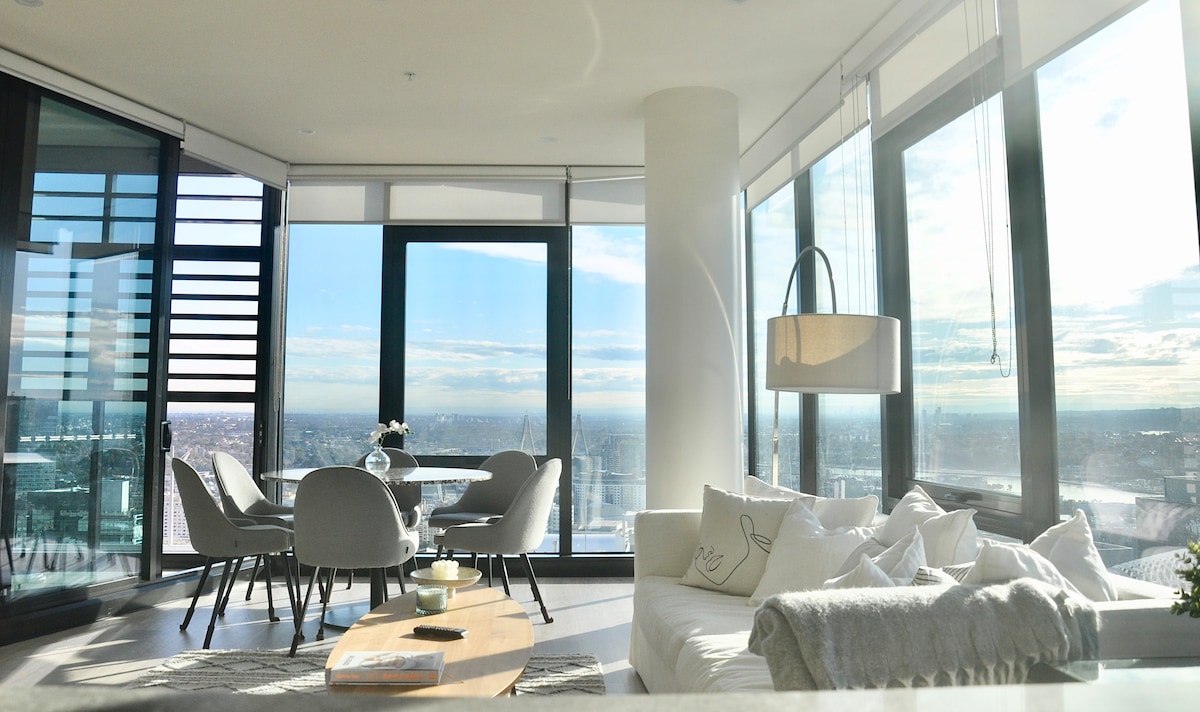 A bright living area is filled with natural light, featuring floor-to-ceiling windows that offer expansive city views. A cozy sofa is paired with a light wooden coffee table, and a dining set with four chairs is positioned near the windows, inviting gatherings.