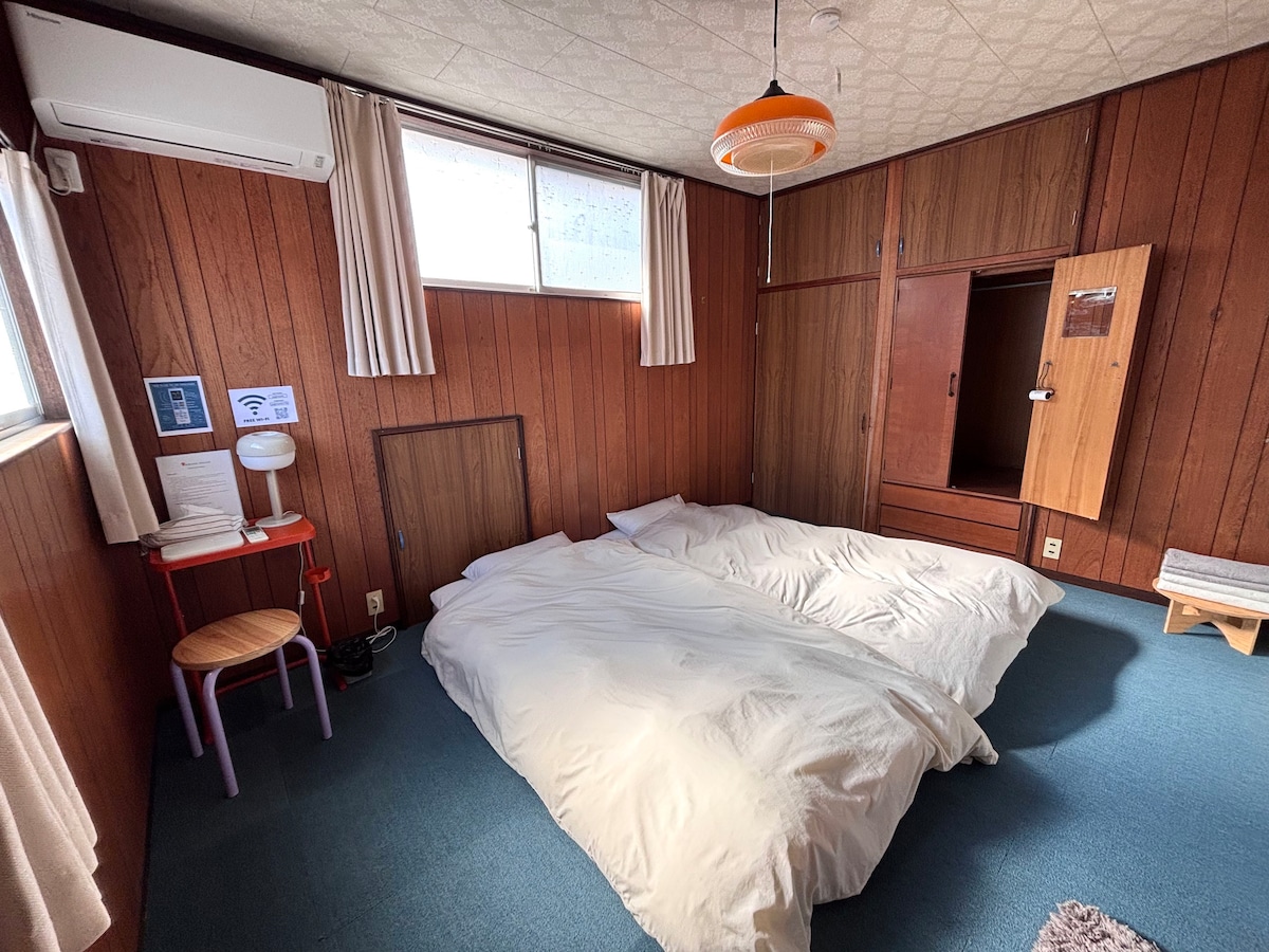 A cozy guest room features two spacious beds dressed in white bedding, set against wooden paneling. Natural light enters through a large window with sheer curtains. A small lamp and a stool are positioned beside the beds, contributing to a simple and functional atmosphere.