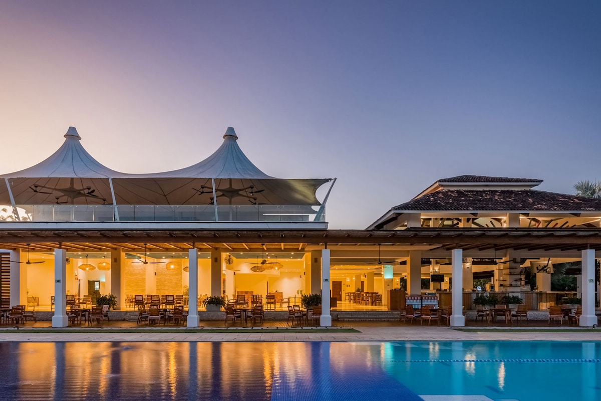 The image captures a stylish poolside restaurant featuring a spacious outdoor dining area under elegant tents. The setting is enhanced by warm lighting reflecting on the water, creating a soothing ambiance. Sleek architecture and lush greenery frame the scene, inviting relaxation and dining enjoyment.