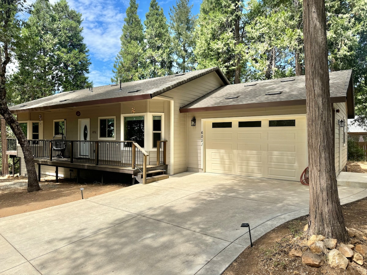 A single-story house is shown with a spacious front porch and a level driveway. The exterior features light-colored siding and a gray roof. Large windows provide natural light, while surrounding tall trees add to the serene environment.