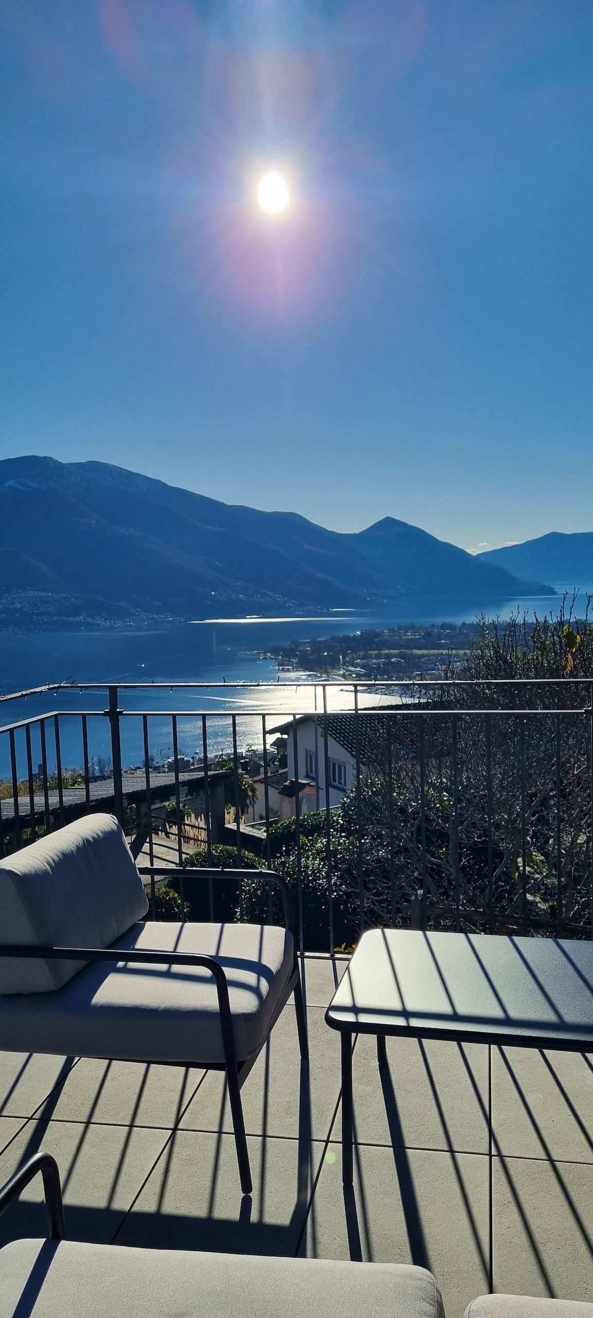 A spacious terrace is depicted, featuring a table and two chairs, offering a clear view of Lago Maggiore and surrounding mountains. The bright sun reflects on the water, illuminating the landscape under a blue sky, creating an inviting outdoor space.