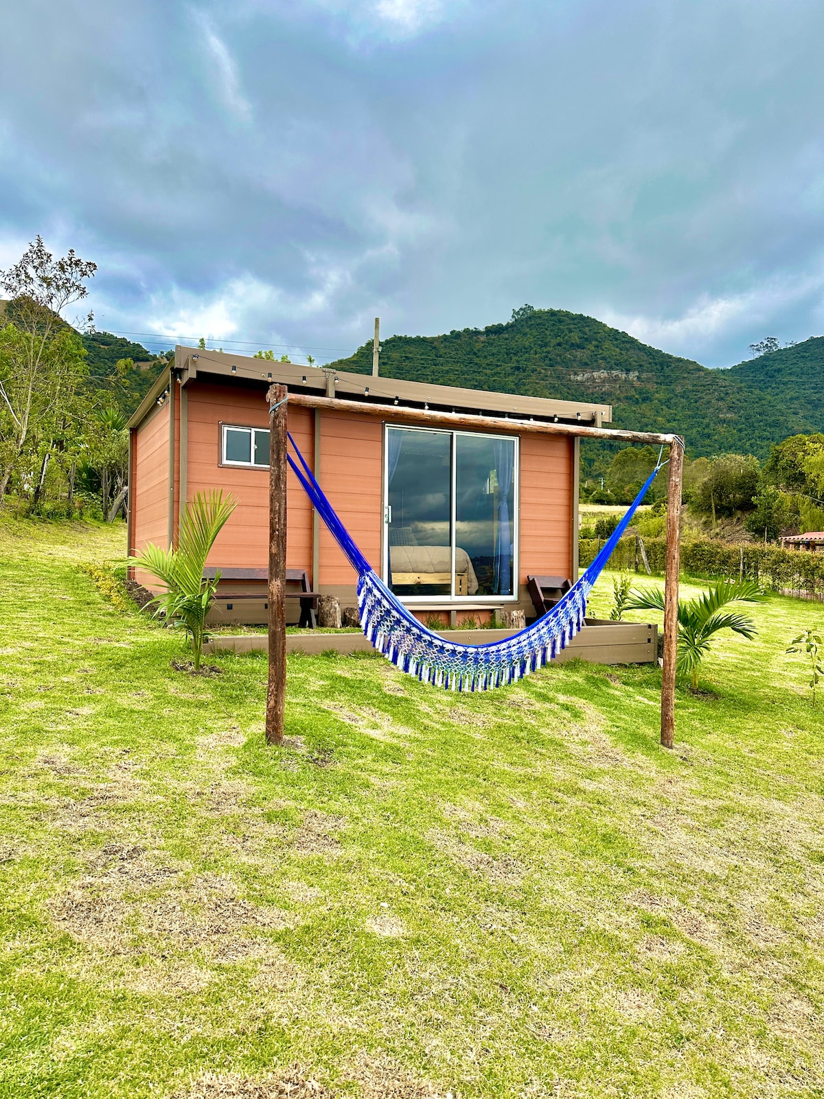 A cabin is positioned on a grassy landscape, framed by lush mountains in the background. A colorful hammock is suspended between two wooden posts, inviting relaxation. Large windows of the cabin reflect the surrounding natural beauty and provide a glimpse of the vibrant greenery.