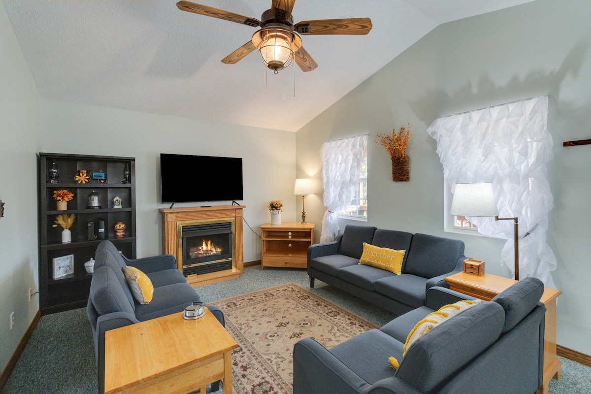 A cozy living room features two dark sofas and a wooden coffee table arranged around a light area rug. A fireplace adds warmth, while large windows allow natural light to fill the space, highlighted by sheer white curtains. A TV is mounted on the wall, providing entertainment options.