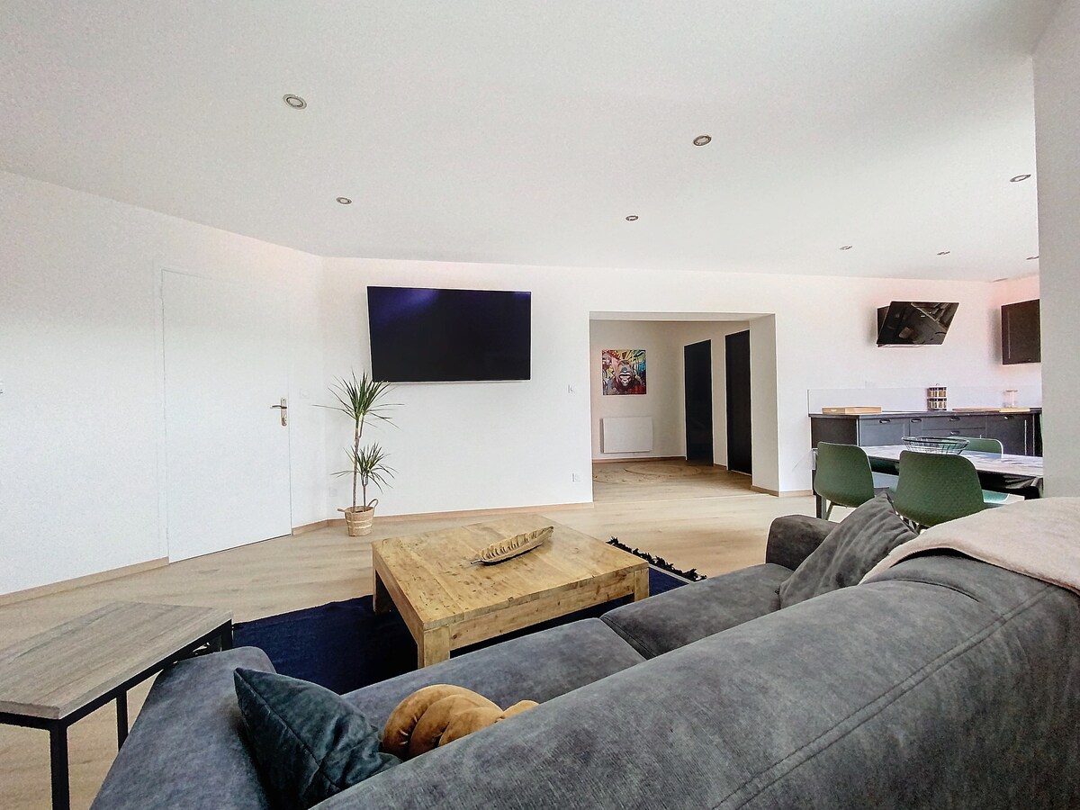 A spacious living area features a large, modern sofa and a wooden coffee table. A wall-mounted smart TV is prominently displayed. The space is well-lit with natural light and connects to a dining area and kitchen in the background, creating an inviting open concept layout.