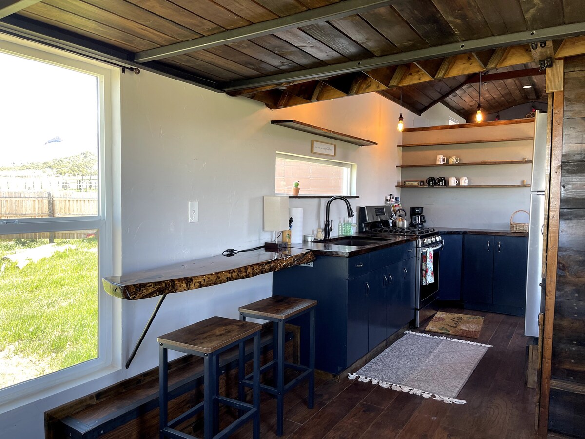 The kitchen features a spacious countertop with natural wood grain, paired with three wooden stools. Modern dark cabinetry complements a large gas stove and oven, along with a sink. Shelves display various mugs and kitchen essentials, while a window provides natural light and views of the outside.
