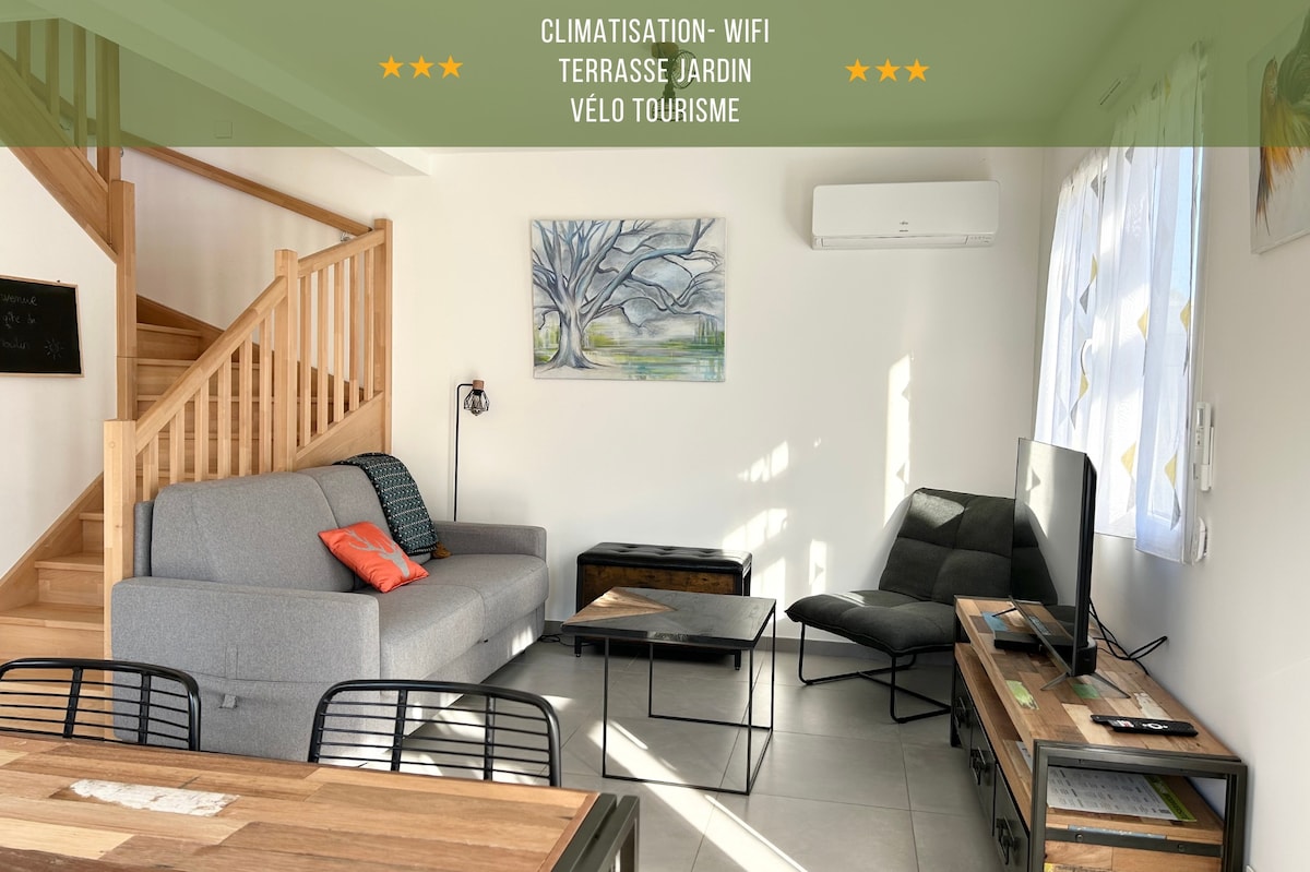 The living area features a cozy gray sofa, accompanied by a modern accent chair and a wooden coffee table. A TV is positioned on a minimalistic stand, while natural light enters through large windows framed with light curtains. A staircase leads to upper levels.