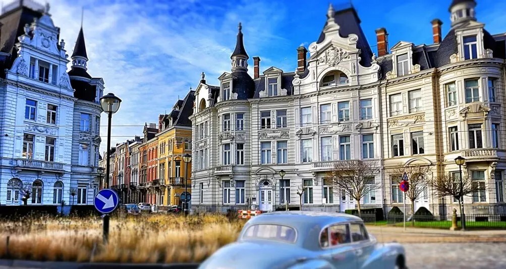 A row of elegant buildings is visible in a vibrant neighborhood, characterized by architectural details and decorative cornices. The scene captures classic facades with intricate designs, surrounded by blooming greenery and a historical vibe. A classic car is parked nearby, enhancing the urban landscape.