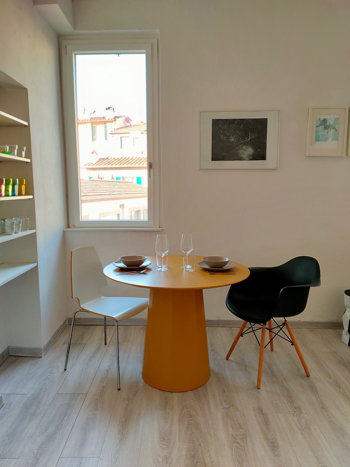 A dining area features a round yellow table paired with one black and one white chair. Table settings include bowls and glasses. A window provides natural light and reveals a view of rooftops, while framed artwork adorns the wall nearby.