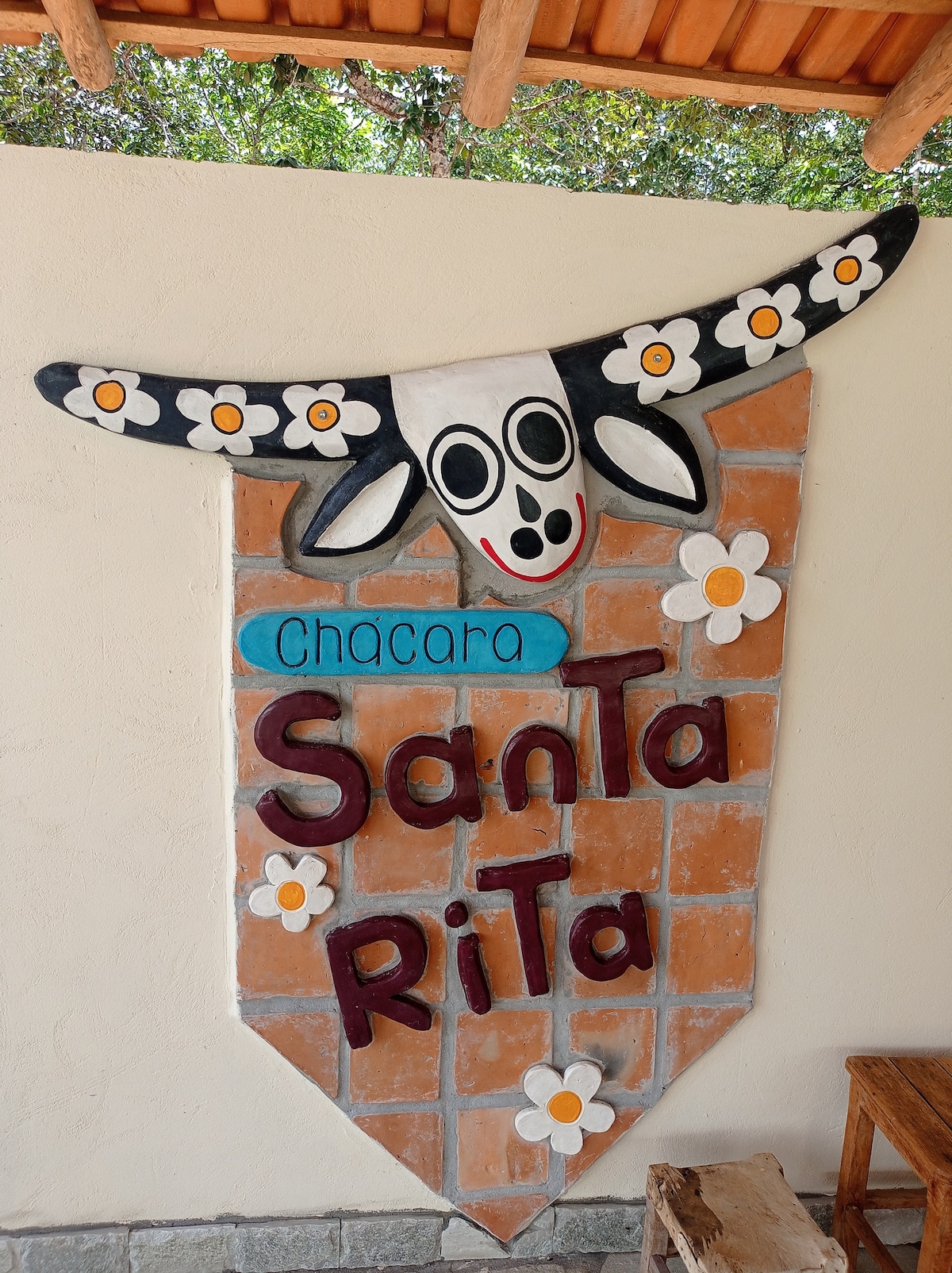 A decorative wall sign displays the name 'Chácara Santa Rita,' featuring a painted cow's head with flowers adornment. The sign is set against a terracotta tile background, conveying a welcoming atmosphere. Natural light filters through foliage, enhancing the view of the entrance.