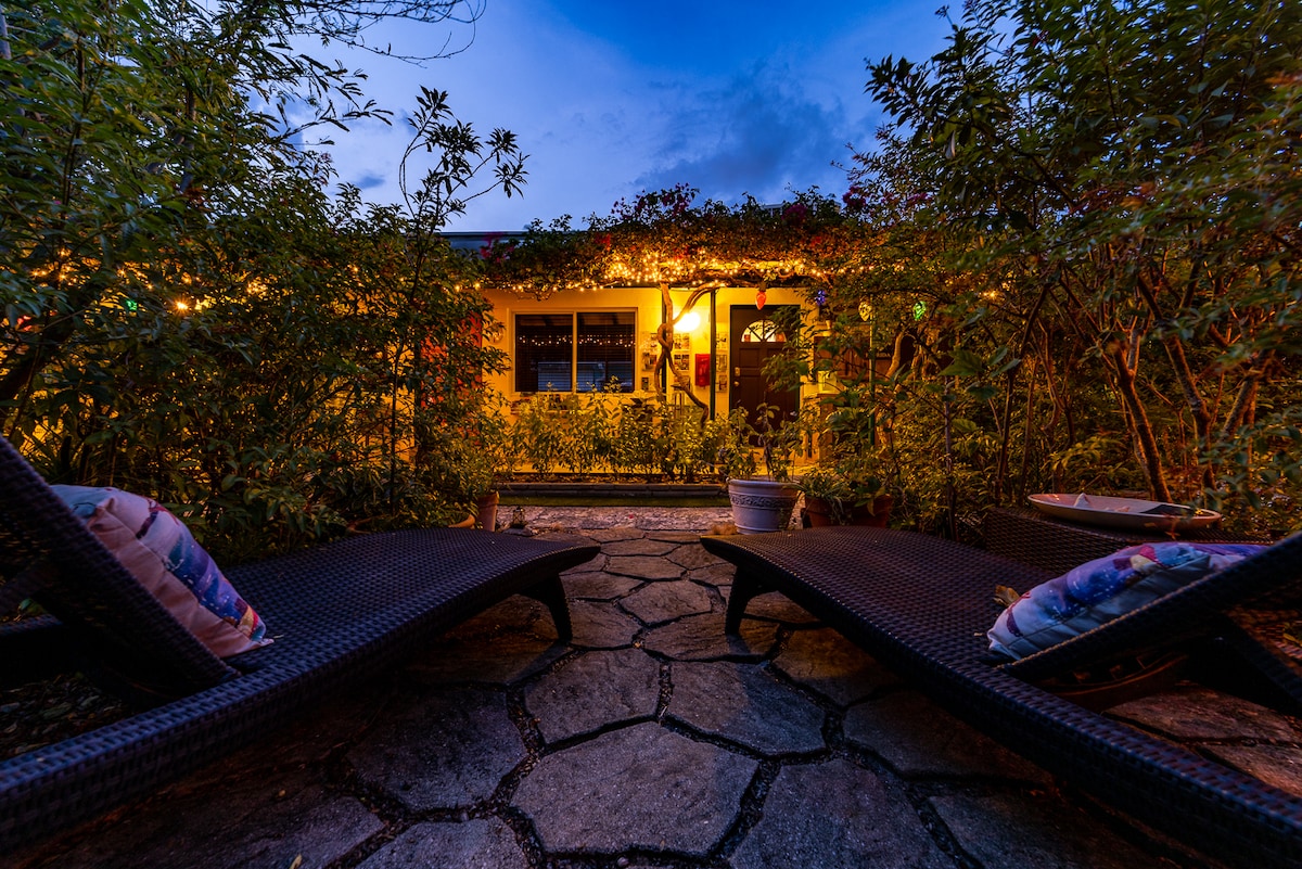 Two lounge chairs with cushions are positioned on a stone pathway that leads towards a warmly lit apartment. Lush greenery surrounds the area, offering a sense of seclusion and tranquility. The evening sky glows softly, creating an inviting atmosphere.