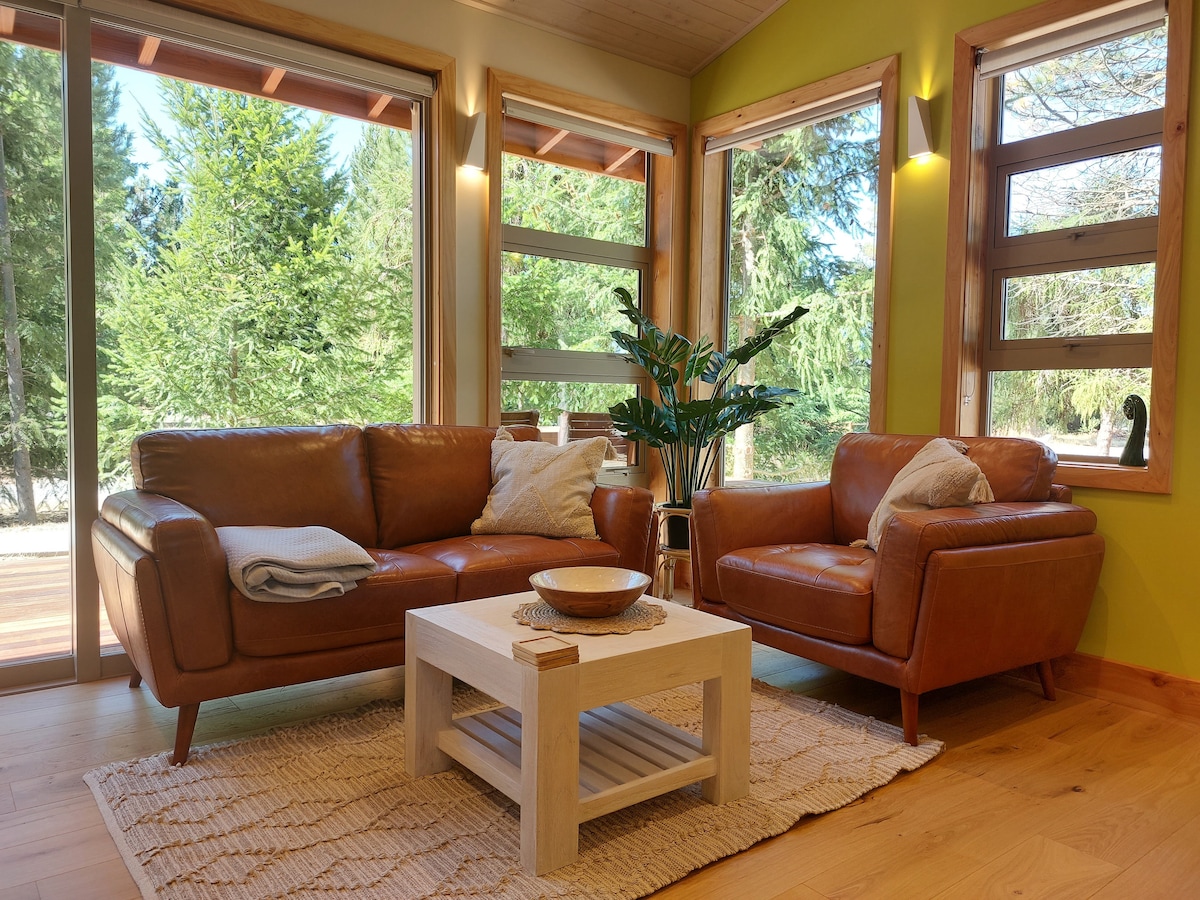 A cozy lounge area features two leather sofas and a central wooden coffee table on a textured rug. Large windows provide views of the surrounding greenery, allowing natural light to brighten the space.