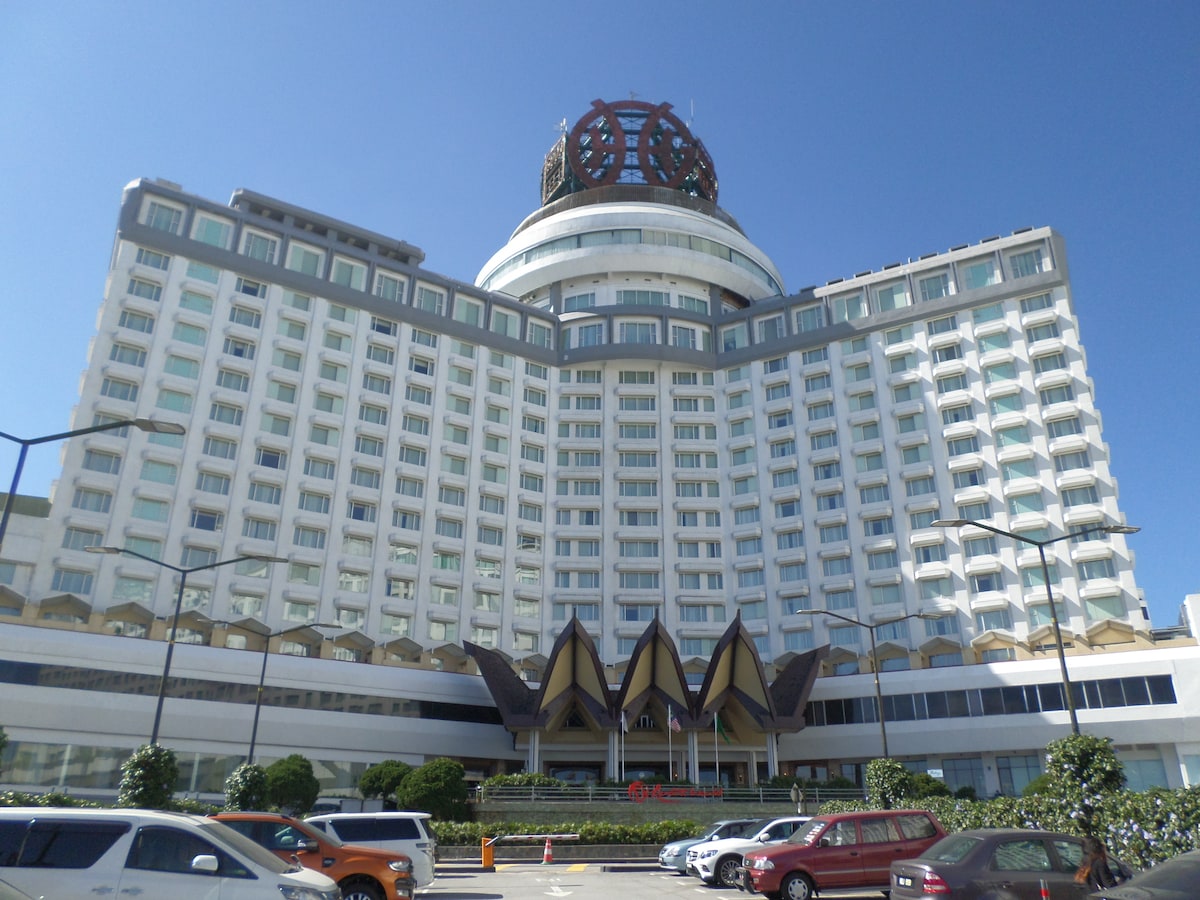 A large hotel building is shown, featuring an intricate and modern architectural design. The facade includes numerous windows and a distinctive rooftop structure. Lush greenery is visible in the foreground, and multiple parked vehicles are positioned along the driveway.