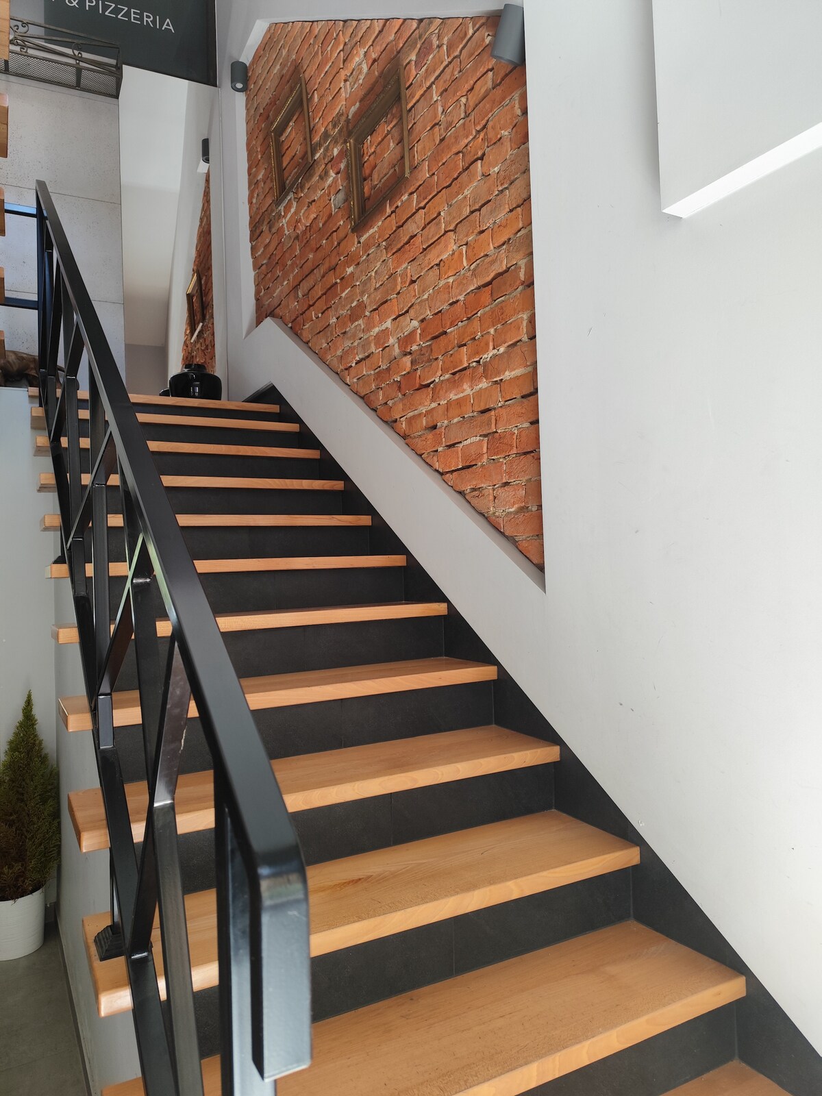 A staircase is depicted, featuring wooden steps and a sleek black railing. The walls showcase a modern design with exposed brick on one side, providing an industrial touch to the space. Potted plants are visible at the base, enhancing the overall ambiance.