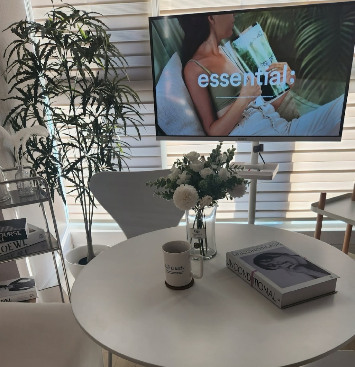 A modern living space is depicted, featuring a round white table at the center. A stylish television offers a relaxing ambiance with a displayed text. Fresh flowers are arranged in a vase, and a stack of books is present, adding to the inviting decor.