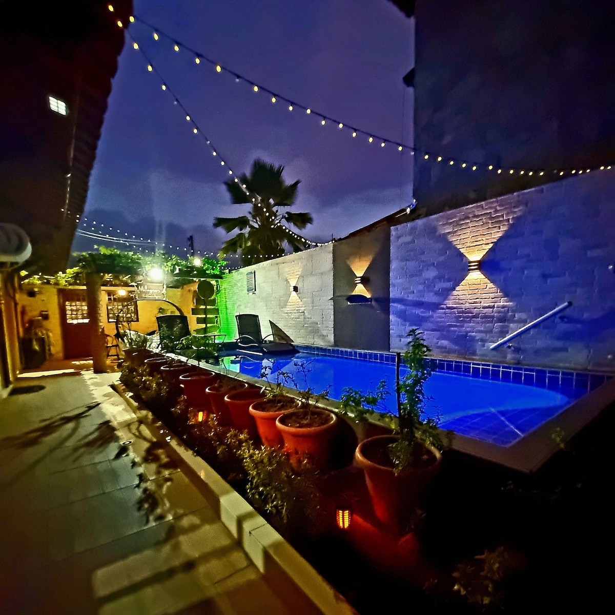 The outdoor area features a swimming pool surrounded by landscaped gardens with potted plants. Soft lighting illuminates the space, reflecting off the water's surface. String lights hang above, creating a pleasant ambiance in the evening setting.