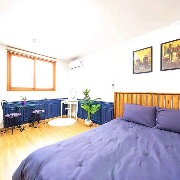 A serene sleeping area is presented with a spacious bed dressed in soft purple bedding. A large window allows natural light to illuminate the room, while a small seating area with a round table and two chairs is visible. Decorative plants and framed artwork add subtle character.