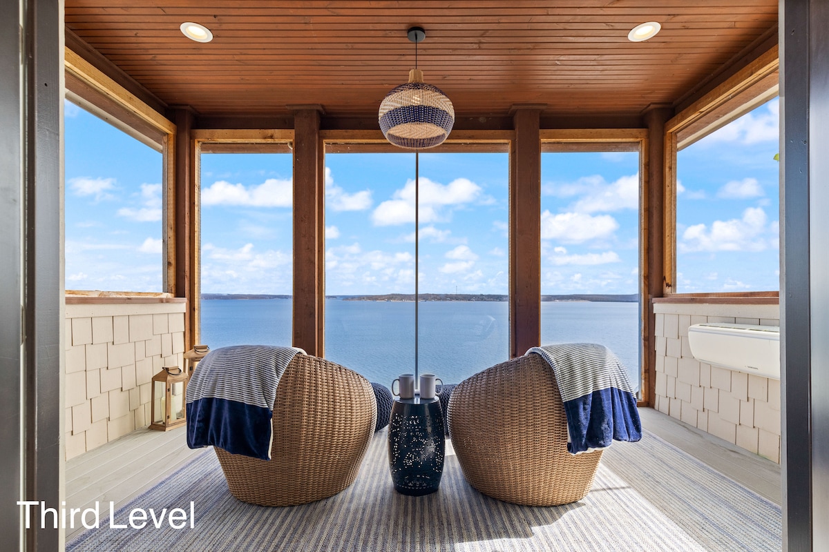 Two wicker chairs with blue blankets are positioned on a balcony, offering unobstructed views of the expansive lake. A decorative table sits between the chairs, and large windows frame the serene landscape, allowing natural light to enhance the inviting space.