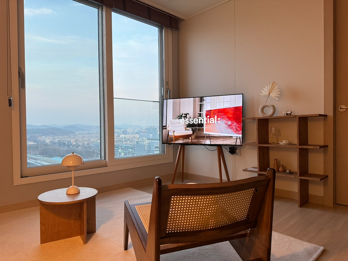 A cozy living area features a sleek wooden chair paired with a round coffee table. A large window offers a view of the city skyline, allowing natural light to fill the space. A modern television is mounted on the wall, adding contemporary flair.
