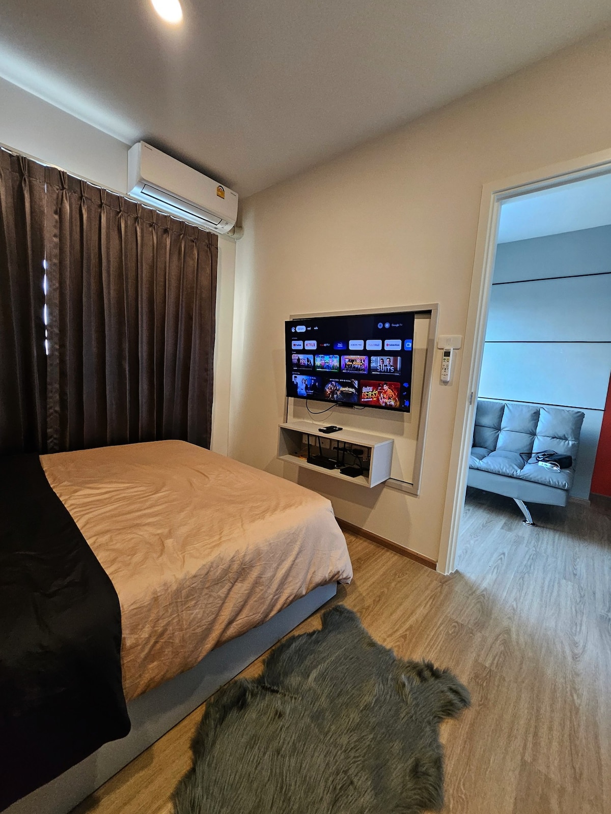 A cozy sleeping area features a spacious bed dressed in a neutral-colored coverlet. A modern television is mounted on the wall opposite. Dark curtains frame a window, while a light area rug adds comfort to the space. A stylish seating area is visible through the doorway.