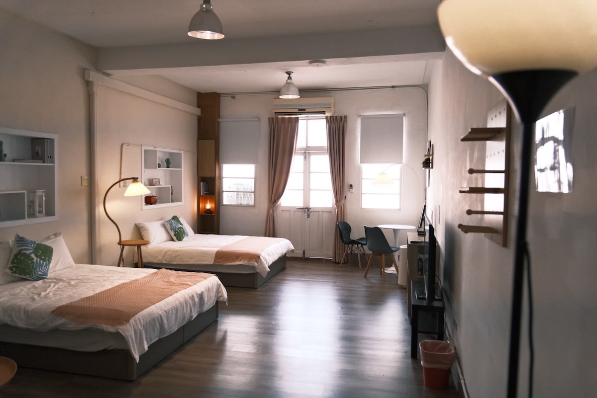 The spacious room features two beds with neutral bedding, each complemented by decorative pillows. Natural light floods in through a large window with light-colored curtains. A comfortable seating area includes blue chairs, while shelves and a lamp add functional decor to the inviting atmosphere.