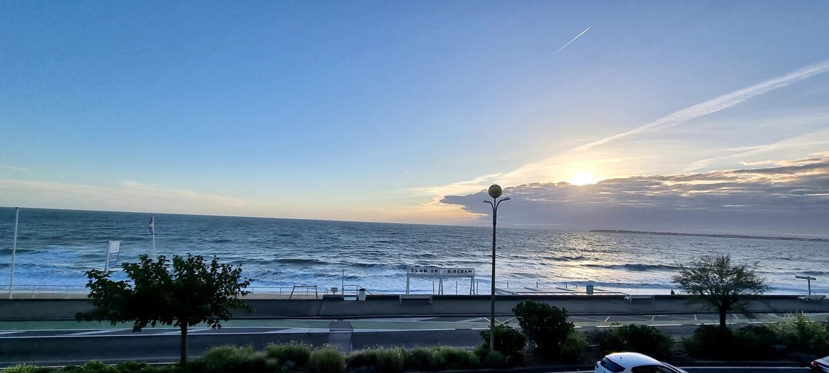 The image presents a coastal view during sunset, showcasing the ocean waves gently rolling towards the shore. A serene sky transitions from blue to orange, while a tree and streetlamps frame the scene, hinting at the beach promenade in the foreground.