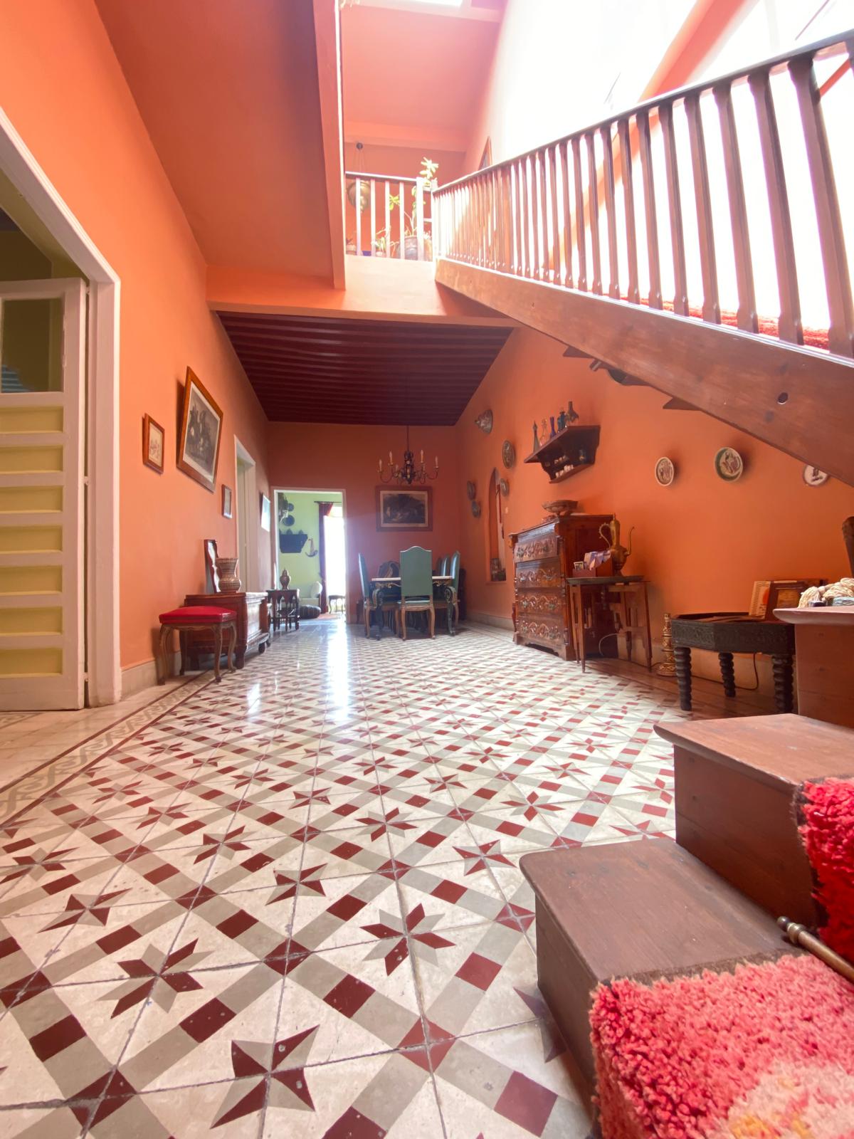 The vibrant hallway features eye-catching patterned tiles that lead to a wooden staircase. Natural light streams in from above, illuminating the warm orange walls. Antique furniture and decorative wall art are visible, enhancing the historic charm of the space.