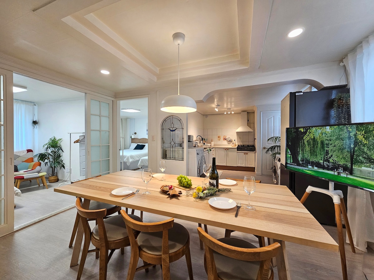 A spacious dining area features a large wooden table set for six, complemented by elegant glassware and a decorative centerpiece. The open layout provides views into the kitchen and living areas, where a comfortable sofa and modern appliances are visible.