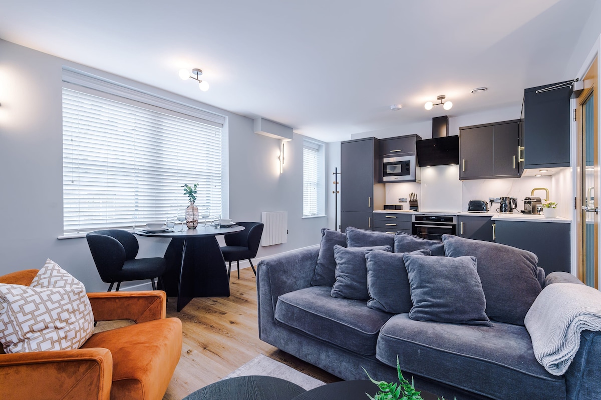 An open-plan living area features a comfortable gray sofa and an orange armchair, offering a cozy seating arrangement. A dining table for four is positioned near a window, while a modern kitchen equipped with sleek appliances is visible in the background.