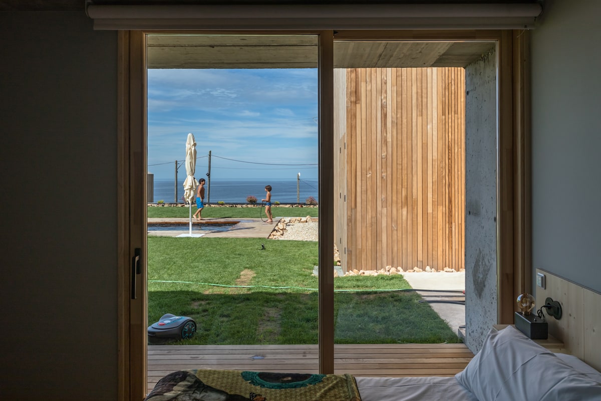 A view from the interior bedroom opens to an outdoor space featuring a lawn, a swimming pool, and a wooden structure in the background. The scene captures individuals enjoying the sun near the pool, with the ocean visible beyond the grassy area.
