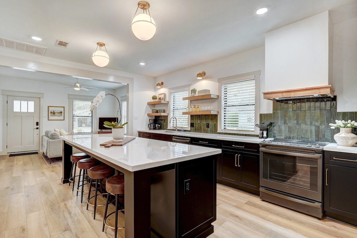 An expansive kitchen is featured, showcasing a large central island with seating for four. High-end appliances and abundant countertop space are highlighted, complemented by warm lighting fixtures and natural light flowing through multiple windows.