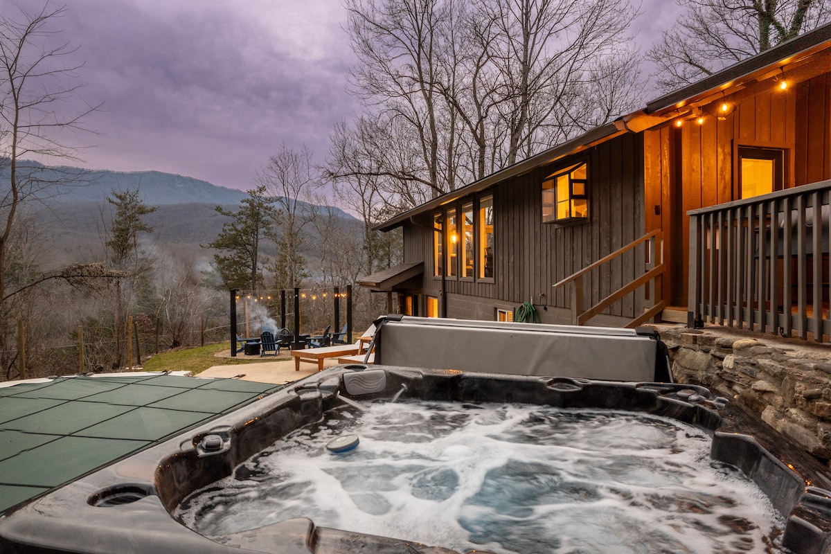 A spacious hot tub is situated on a patio, offering mountain views under a twilight sky. The cabin's exterior is visible, featuring large windows that reflect the surrounding nature. Soft outdoor lighting creates a cozy ambiance around the space.